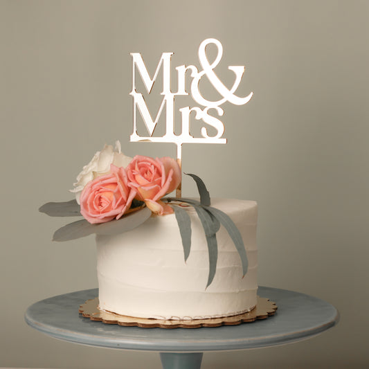 Wedding cake topper in mirror gold acrylic "Mr & Mrs" sitting on a white cake with flowers on top. 