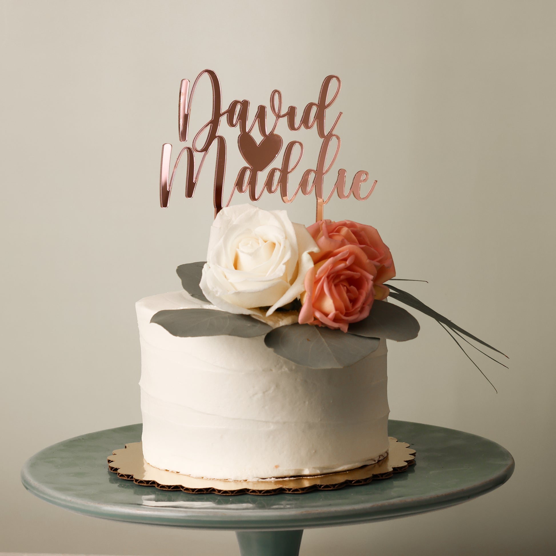 Wedding cake topper in mirror Rose gold acrylic "David Heart Maddie" sitting on a white cake with flowers on top.