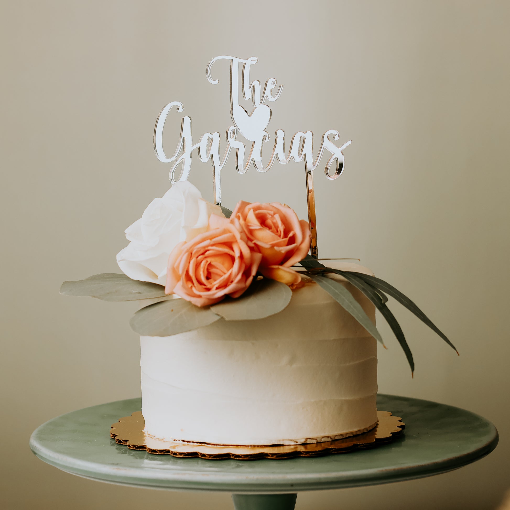 Wedding cake topper in mirror Silver acrylic "The Garcias" sitting on a white cake with flowers on top.